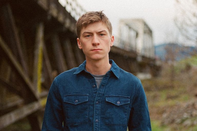 Country music artist Sam Barber wearing a blue denim shirt stands outdoors looking directly at the camera with a bridge in the background.